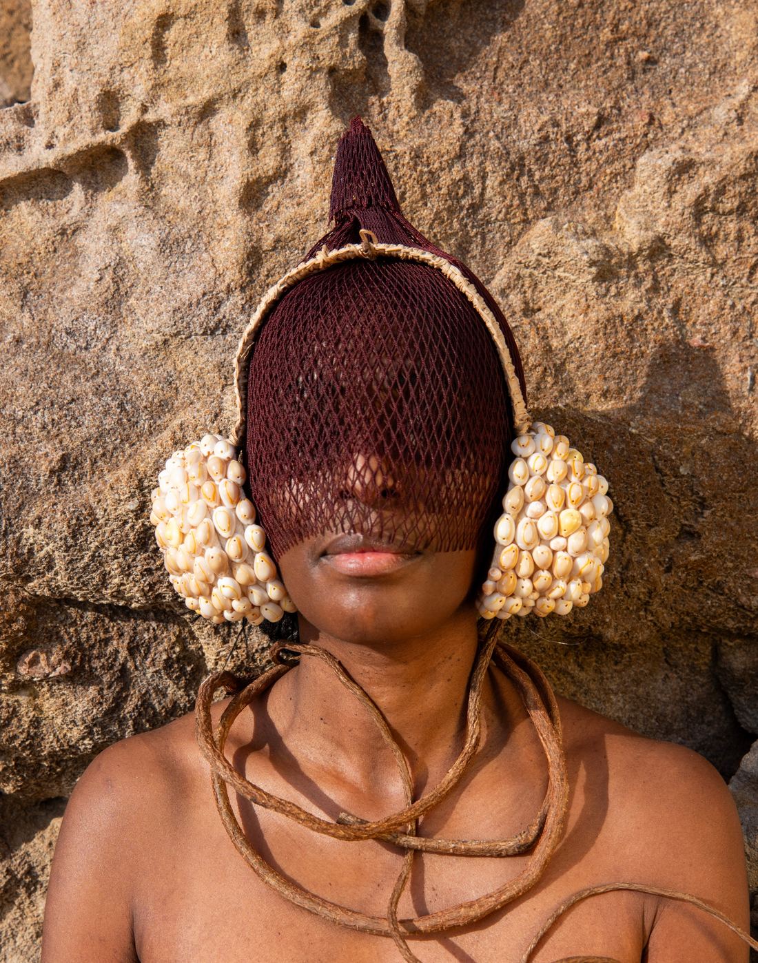 Gros plan du buste : un masque en résille brune couvre le haut du visage et laisse voir la bouche ; deux oreillettes de cauris blancs encadrent la tête ; un cordon de fibre brune s'enroule autour du cou et des épaules nues.