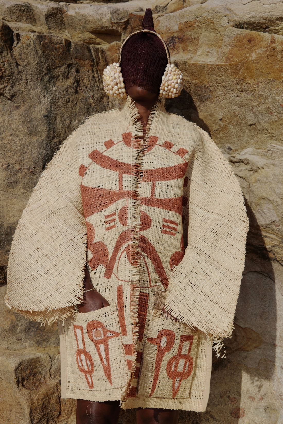 Une femme debout contre une paroi de roche ocre porte un long manteau de raphia écru orné de motifs géométriques terre cuite. Son visage est entièrement couvert d'un masque brun à crête pointue, encadré de deux oreillettes bombées couvertes de cauris.