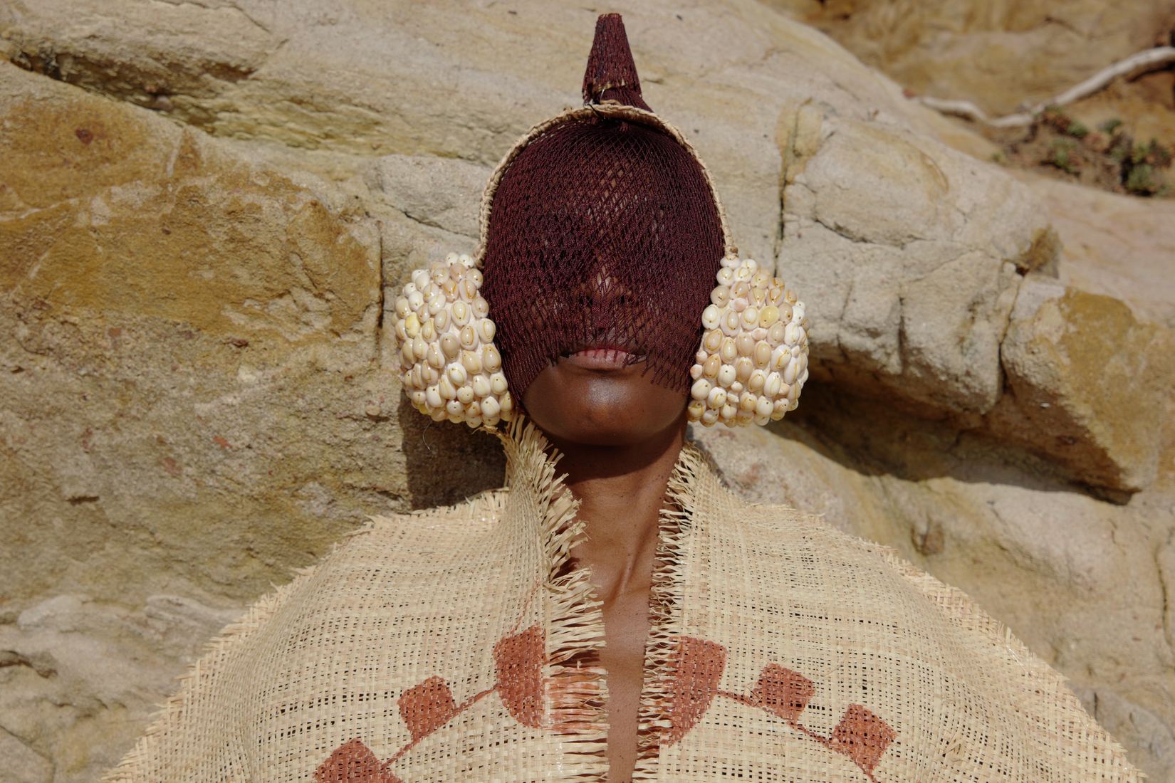 Plan horizontal à mi-corps : la femme masquée est adossée à la roche ocre, le masque brun à crête et les oreillettes de cauris au centre, le haut du manteau de raphia à motifs terre cuite visible au premier plan.