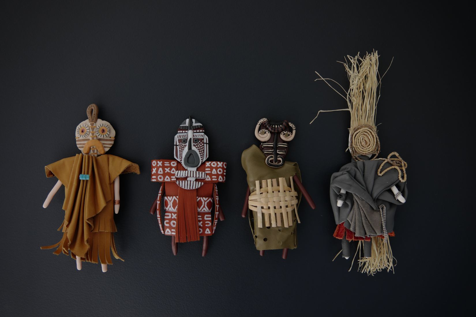 Quatre poupées alignées sur fond noir : vêtues de tissus ocre, rouge, kaki et gris, elles portent des masques peints à motifs géométriques ; la dernière est coiffée d'une gerbe de raphia.