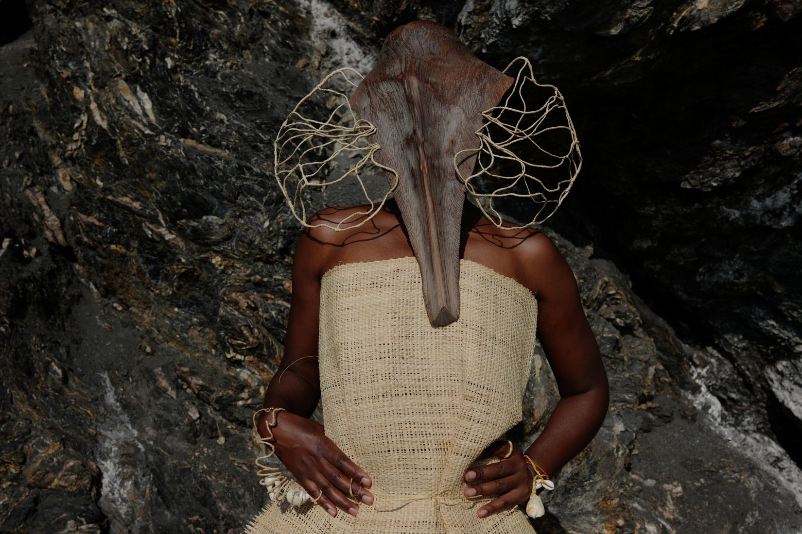 Plan horizontal à mi-corps : devant une roche sombre, une femme aux mains sur les hanches porte un bustier de raphia tissé ; son visage est caché par un long masque brun en forme de bec, entouré d'une auréole de fil de raphia clair.