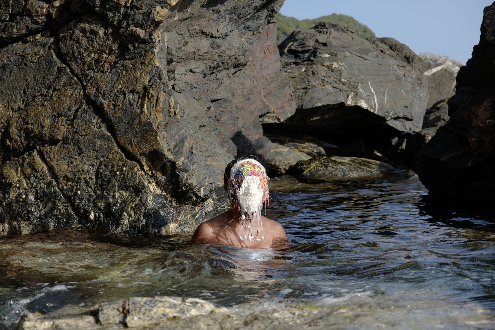 Plan large horizontal : dans une crique entre de hauts rochers, une femme est immergée jusqu'aux épaules dans une eau claire ; son visage est couvert d'un masque blanc de coquillages surmonté de fils colorés.
