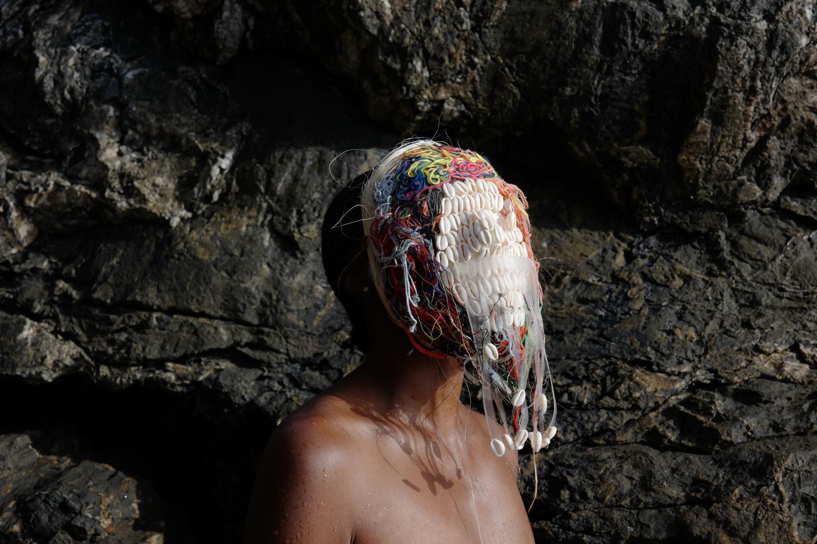Plan horizontal : contre une roche sombre, la femme de profil, épaules nues, porte un masque de cauris blancs dont pendent de longs fils ; le haut du masque est tissé de fils rouges, bleus et jaunes.