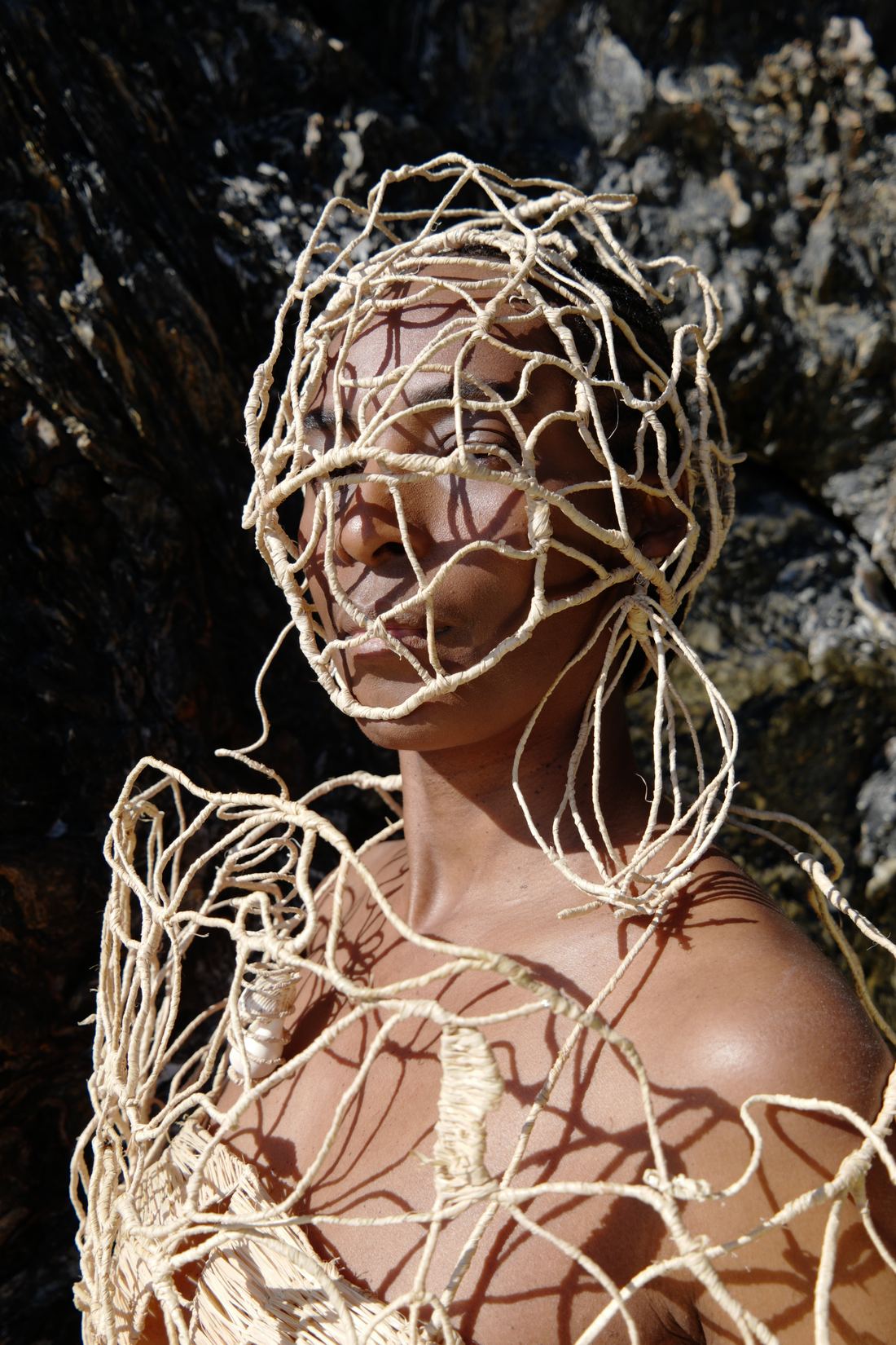 Plan vertical en lumière rasante : le réseau de raphia couvre le visage et descend sur le torse ; le soleil projette l'ombre des mailles sur la peau.