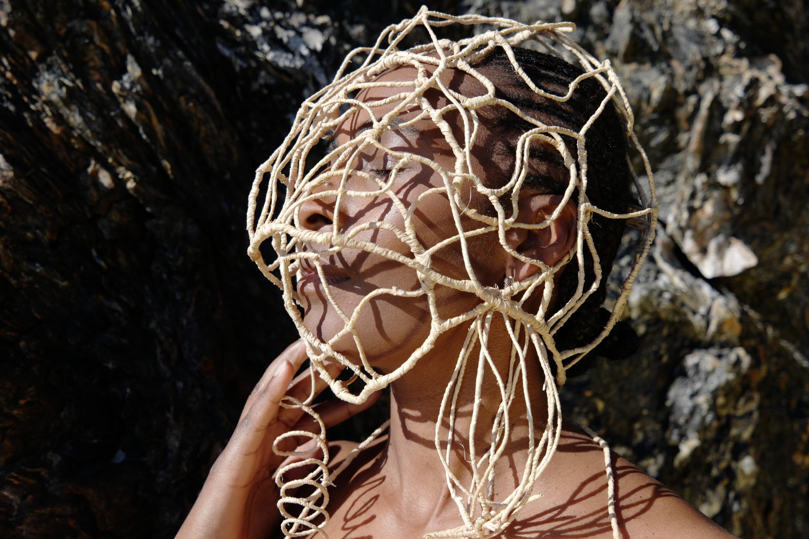 Plan horizontal : tête levée, une main près du visage, la femme porte un masque-cage de raphia qui enveloppe la tête et se prolonge sous le menton ; roche sombre en fond.