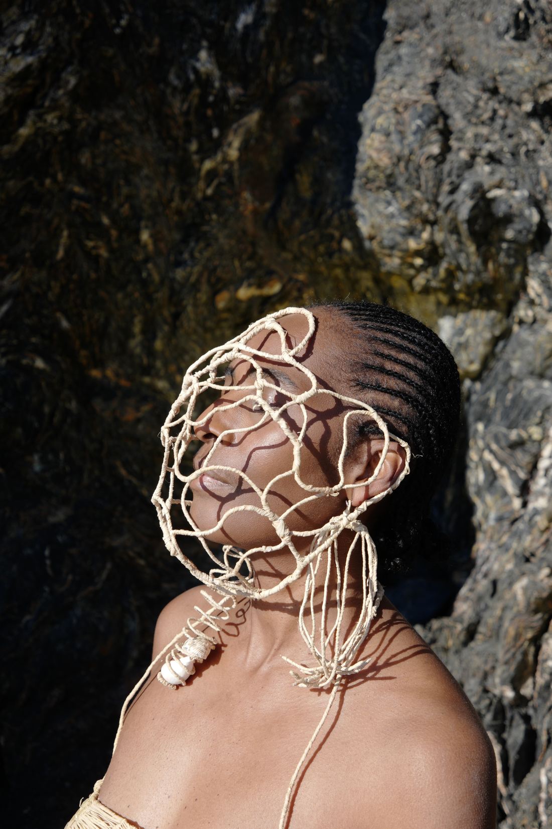 La tête renversée vers le ciel, yeux fermés, la femme porte un masque-cage de raphia qui épouse le visage et se prolonge sous le menton ; cheveux tressés, roche sombre en fond.