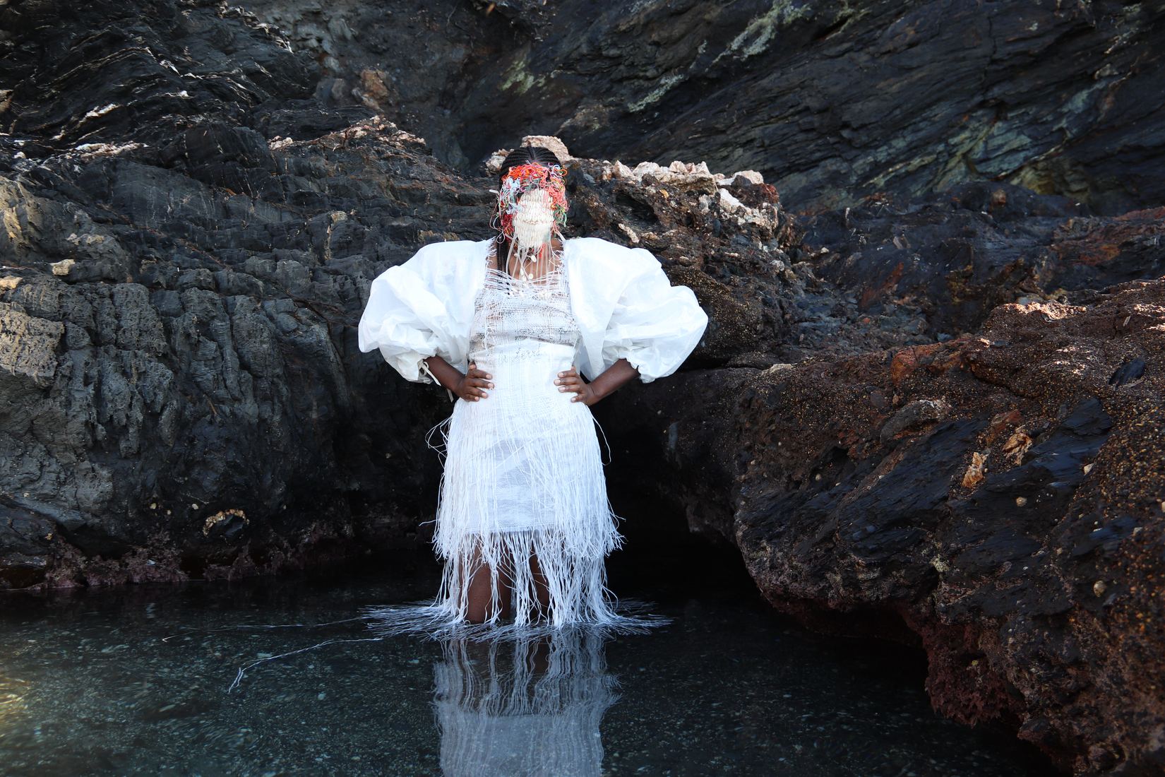 Plan large horizontal : dans une grotte marine, une femme debout dans l'eau peu profonde, mains sur les hanches, porte une robe blanche à grandes manches bouffantes et longues franges ; son reflet apparaît dans l'eau.