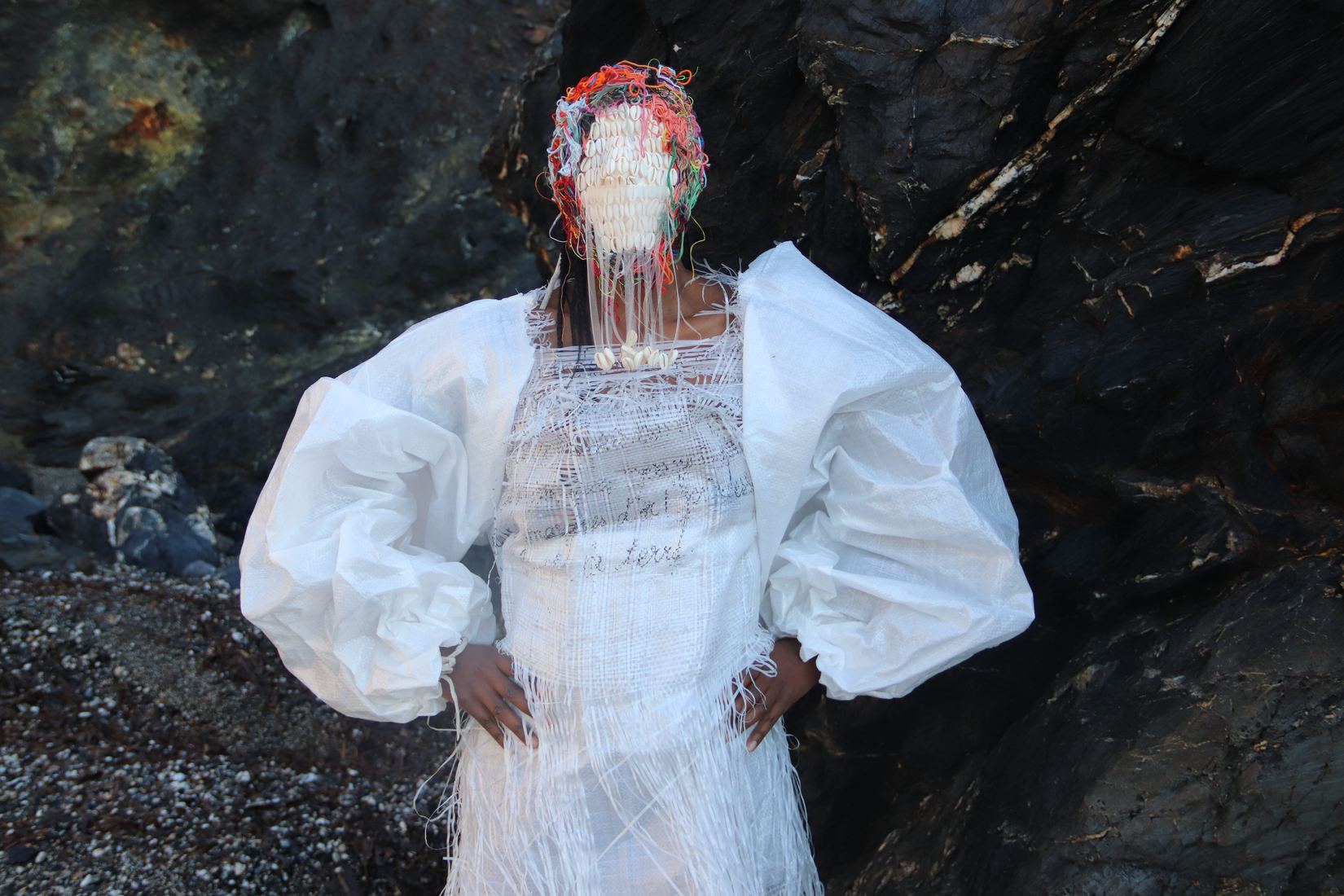 Plan horizontal à mi-corps : la même robe blanche contre la roche sombre ; le visage est couvert d'un masque blanc de coquillages surmonté de fils rouges.