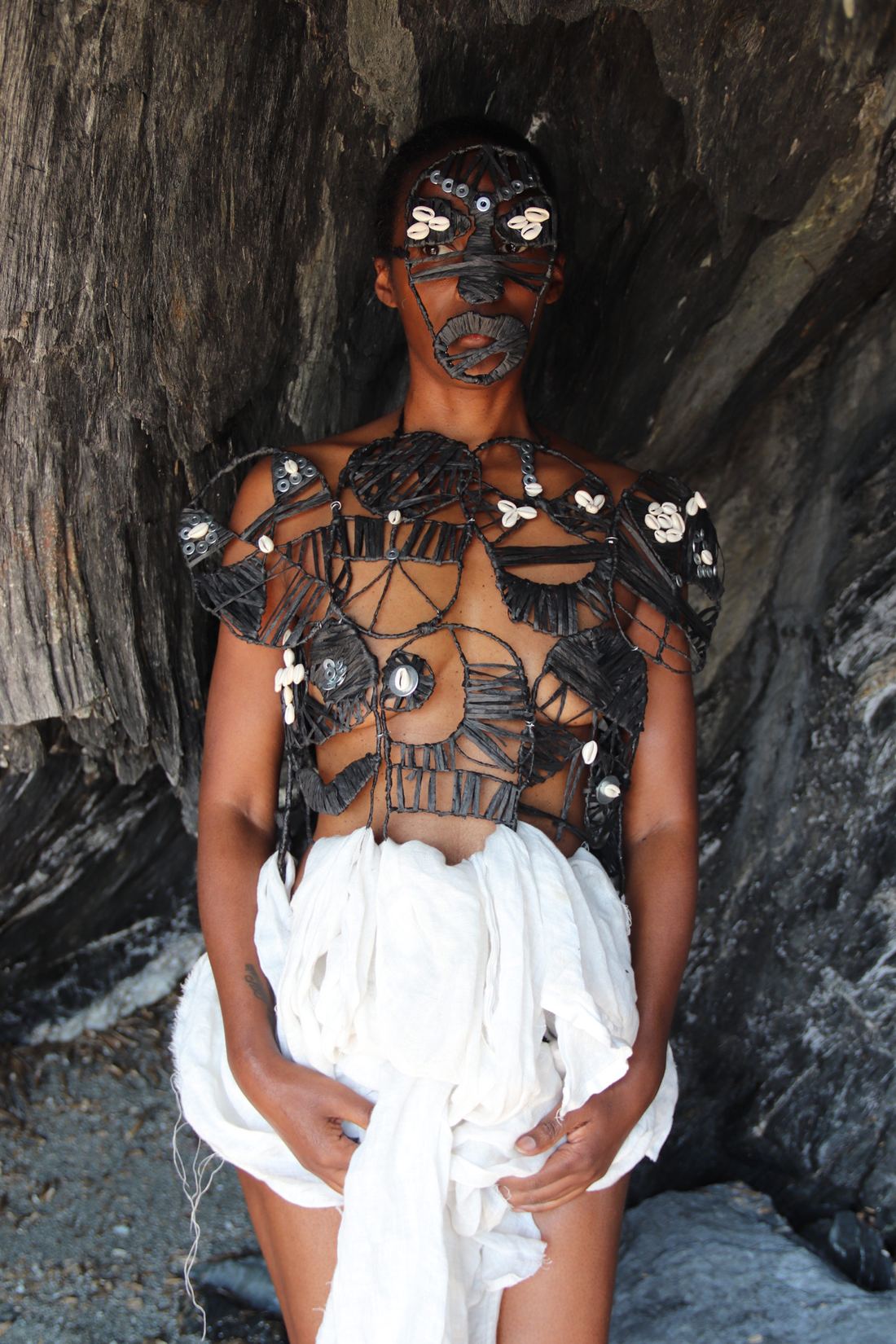 Plan vertical en pied : dans une grotte, une femme debout porte un plastron noir ajouré orné de cauris et un masque noir assorti ; un tissu blanc drapé autour des hanches.