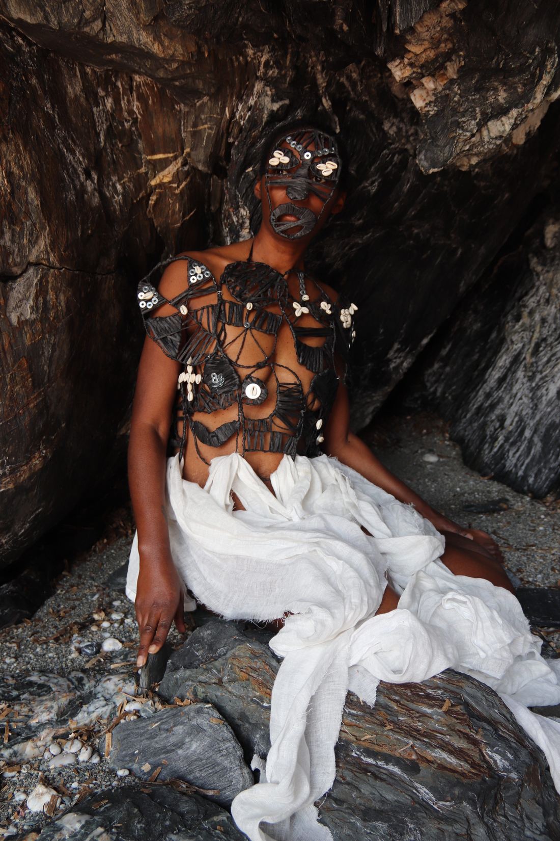 La femme est assise sur un rocher, le tissu blanc étalé autour d'elle ; plastron noir et masque à cauris.