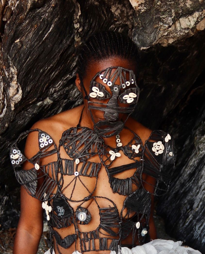 Gros plan du buste : la structure noire tressée du plastron, constellée de cauris et de boutons blancs ; le masque couvre le bas du visage et les yeux.
