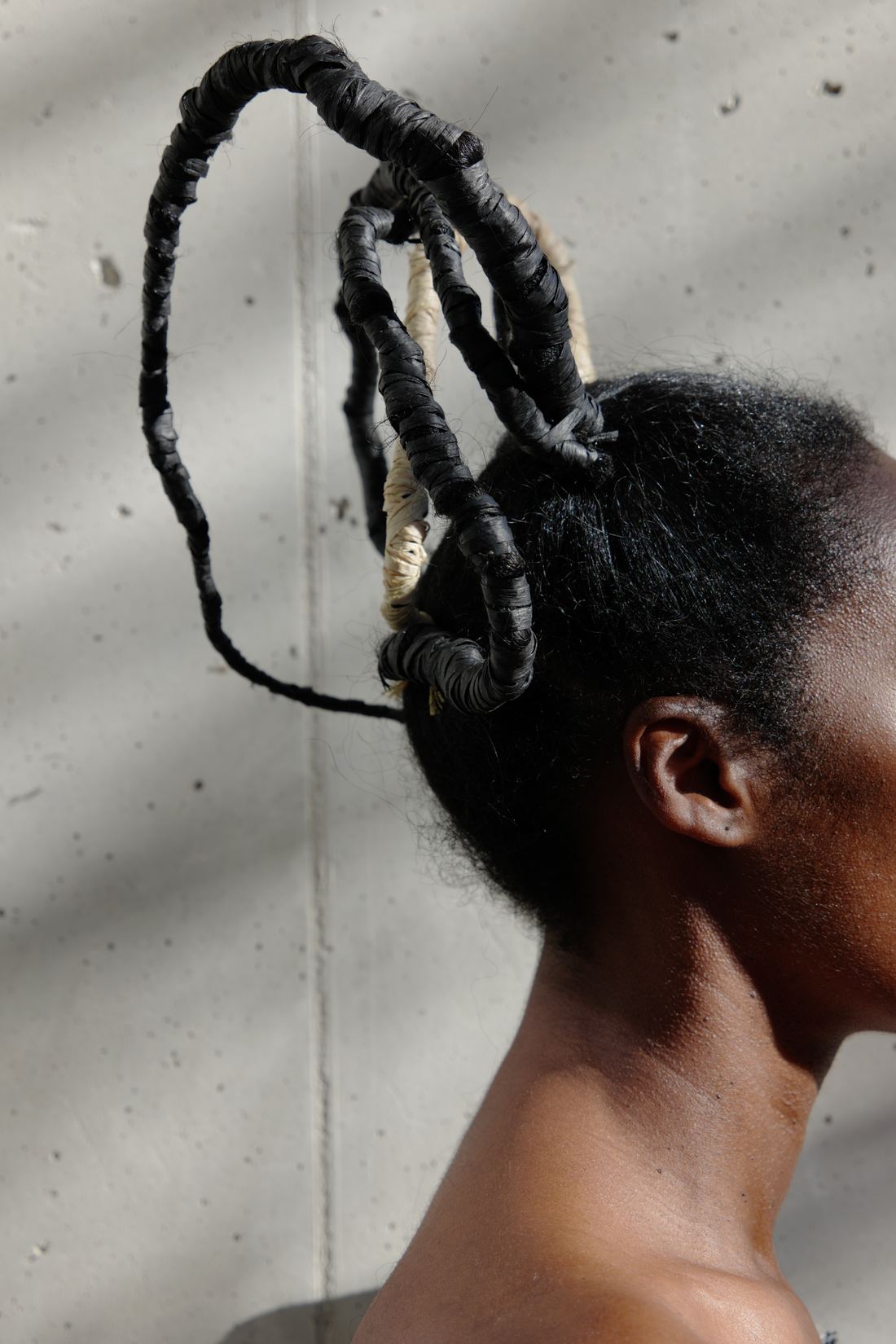 Plan vertical de profil : une femme porte dans ses cheveux une sculpture de grandes boucles noires gainées, avec des segments de raphia clair, devant un mur de béton.
