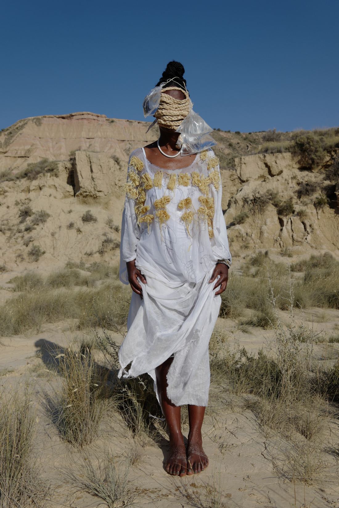 Plan vertical en pied : dans un paysage désertique de collines arides sous un ciel bleu, une femme pieds nus en robe blanche brodée de raphia ; son visage est couvert de sachets d'eau et de raphia.