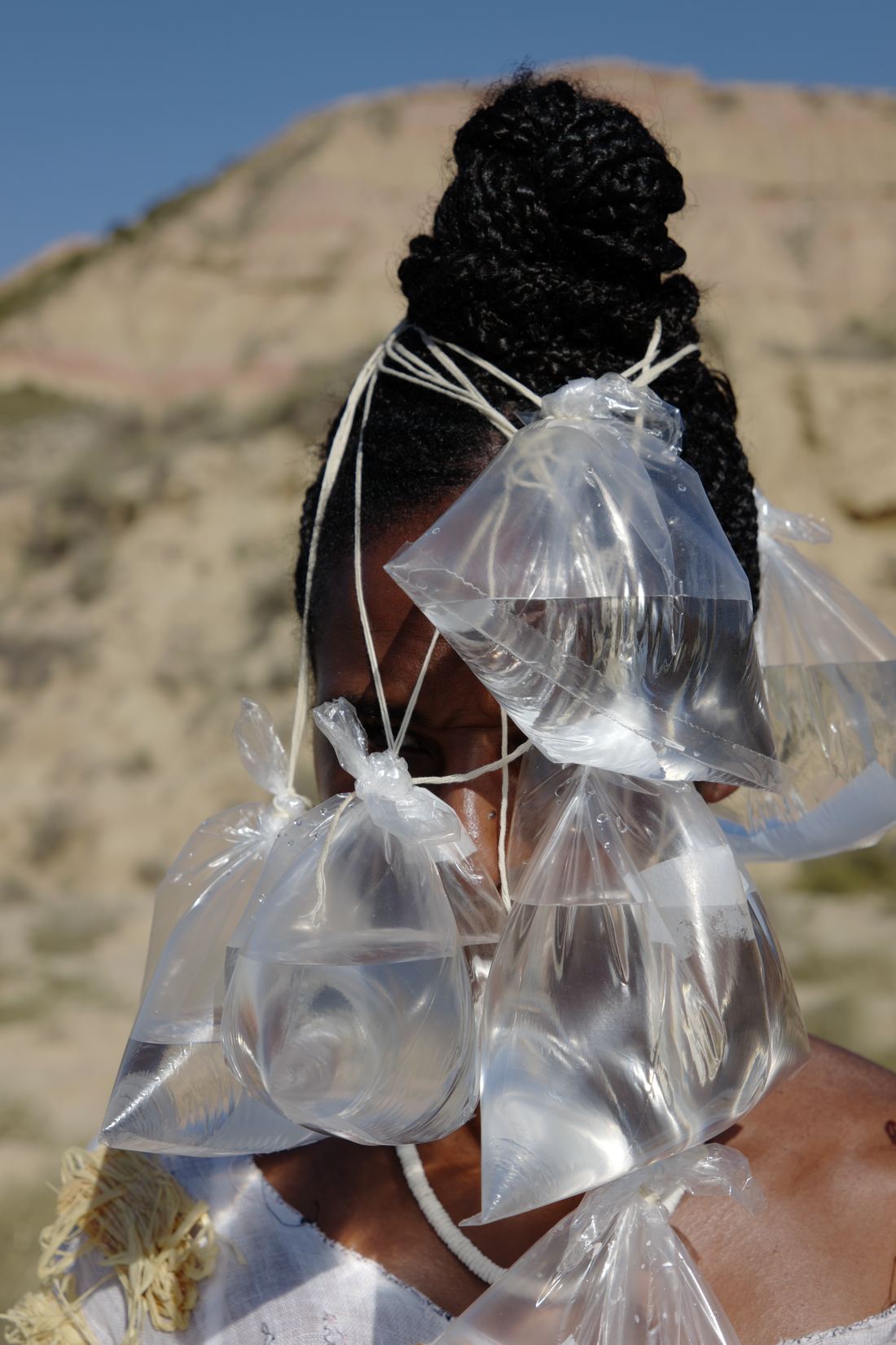 Gros plan : plusieurs sachets transparents remplis d'eau, noués, couvrent le visage ; chignon tressé, colline aride en fond.