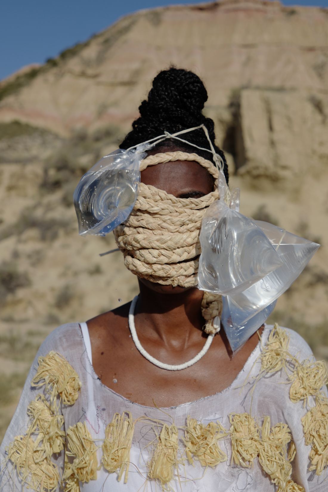 Plan rapproché du buste : un masque de raphia enroulé couvre le bas du visage, des sachets d'eau transparents suspendus de part et d'autre ; collier blanc, broderies de raphia sur la robe.