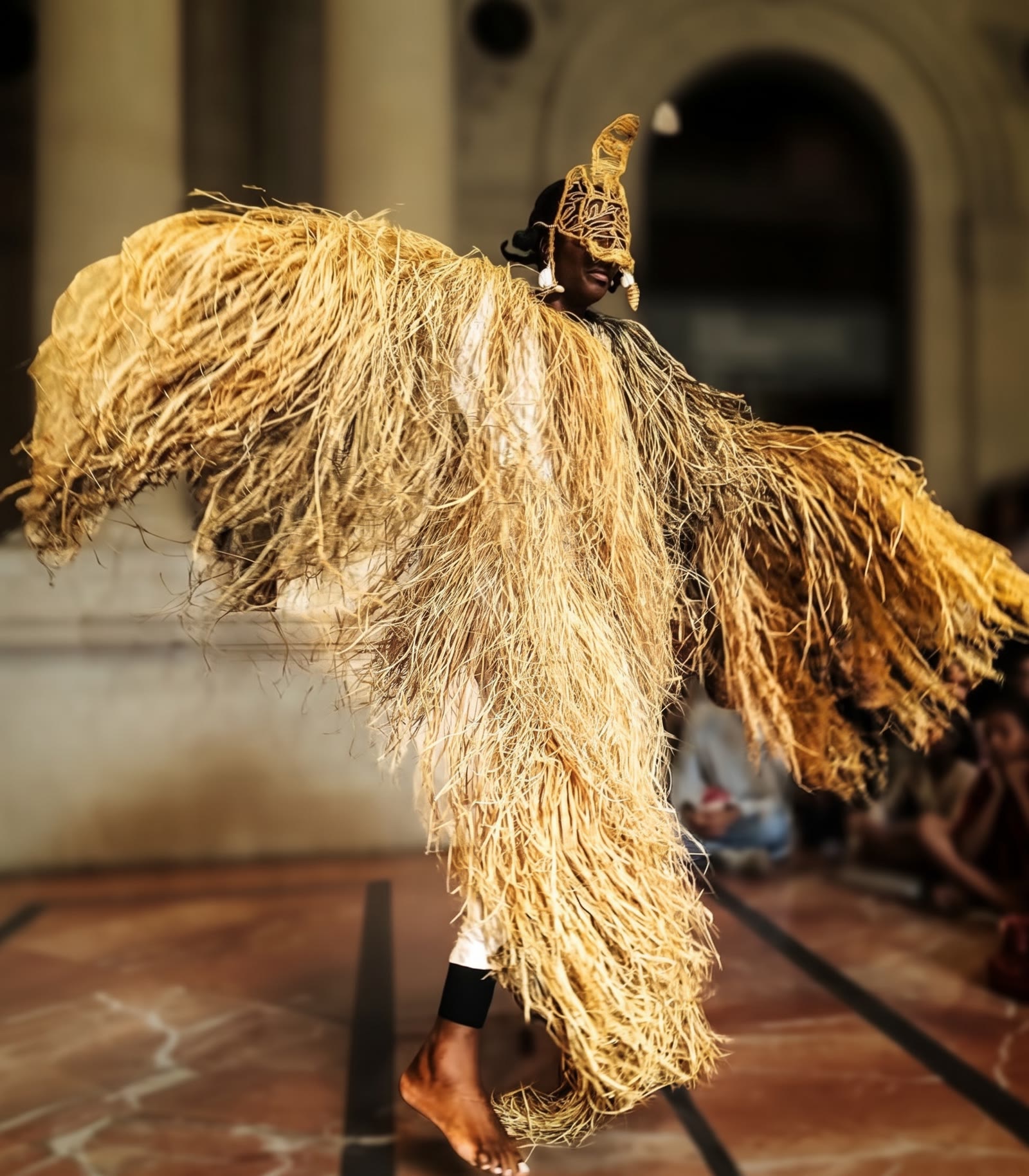 Daja au Musée d’Aquitaine, les bras ouverts, portant un masque et une ample parure de raphia.